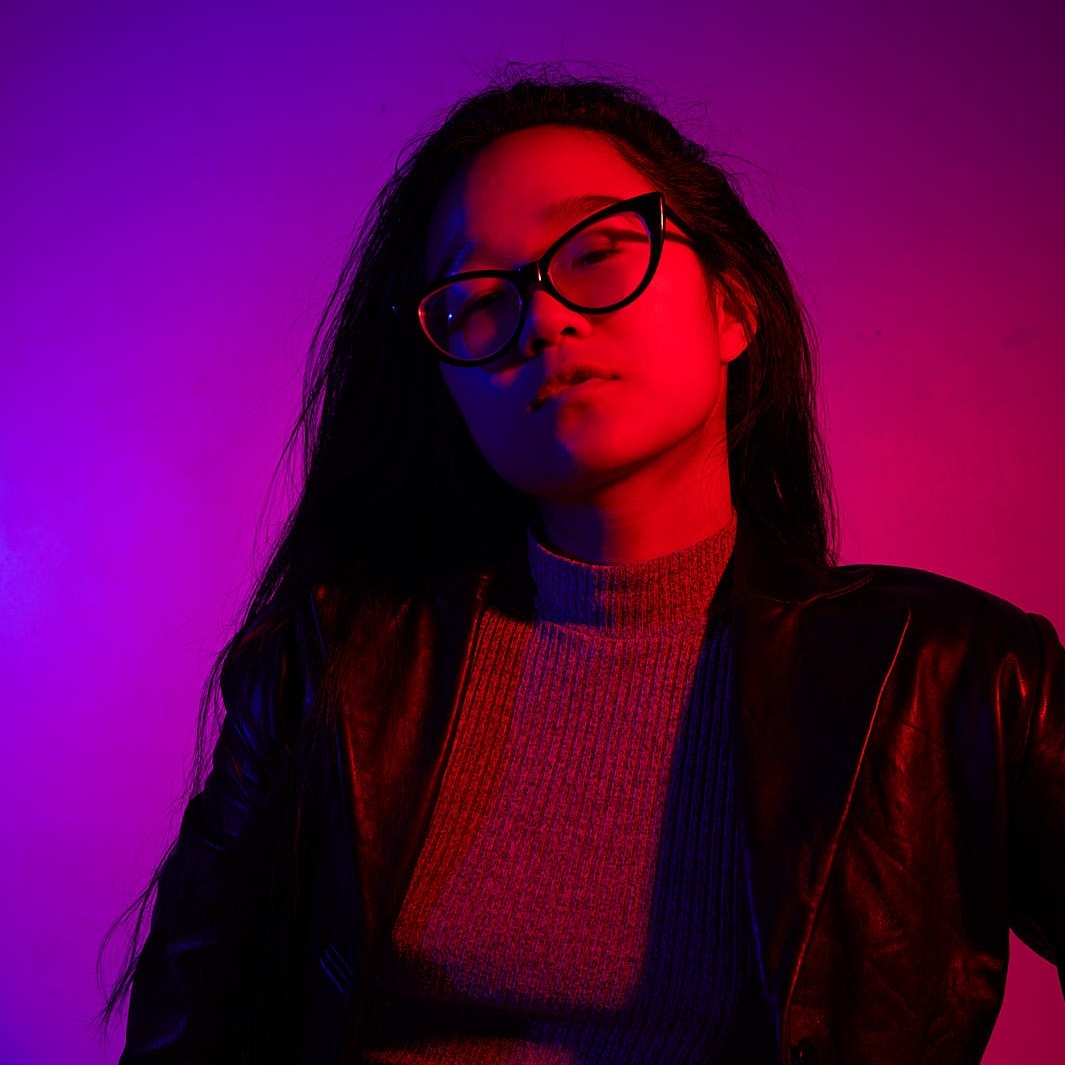 person with long black hair and glasses and black jacket in front of purple and pink backdrop